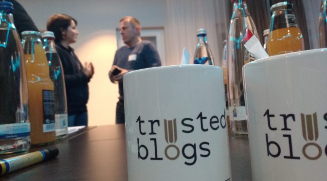 Zwei Tassen mit dem Logo der Firma Trusted Blogs, mit dem Geschäftsführer im Hintergrund