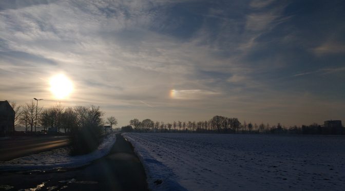 Winterlandschaft mit tiefstehender Sonne und einer kleinen Nebensonne rechts, sieht aus wie ein Regenbogen