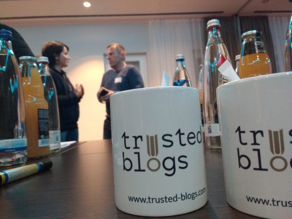 Zwei Tassen mit dem Logo der Firma Trusted Blogs, mit dem Geschäftsführer im Hintergrund