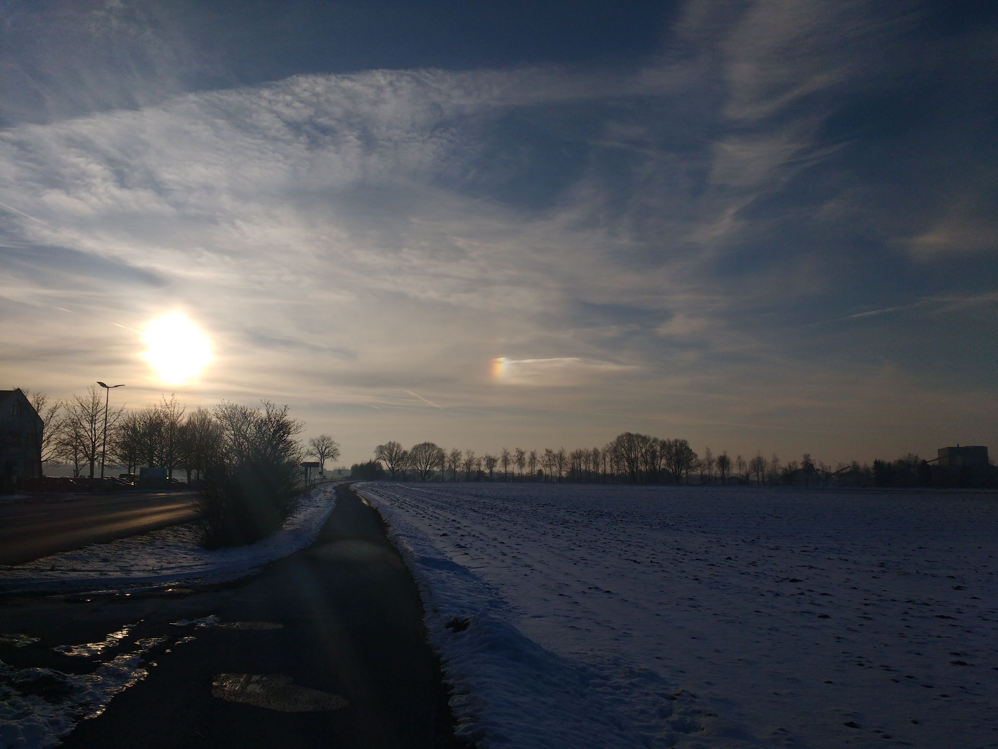 Winterlandschaft mit tiefstehender Sonne und einer kleinen Nebensonne rechts, sieht aus wie ein Regenbogen